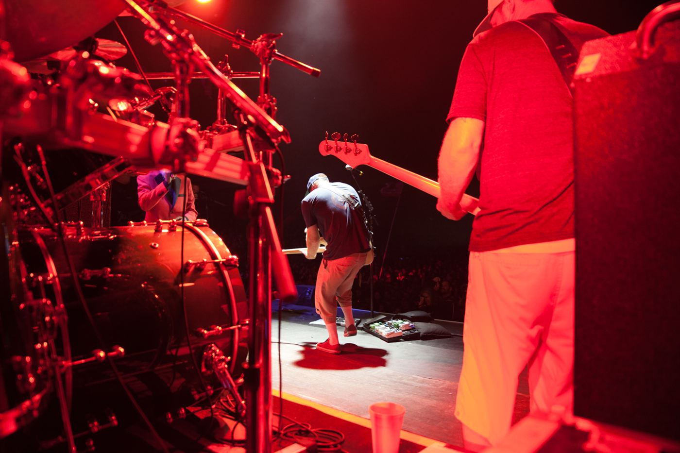 View from the stage behind the drum kit during a live band performance under red stage lighting.